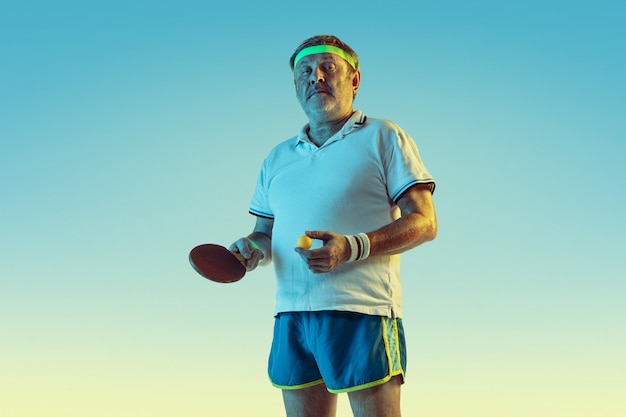 Senior man playing table tennis on gradient wall in neon light. Caucasian male model in great shape stays active, sportive. Concept of sport, activity, movement, wellbeing, healthy lifestyle.