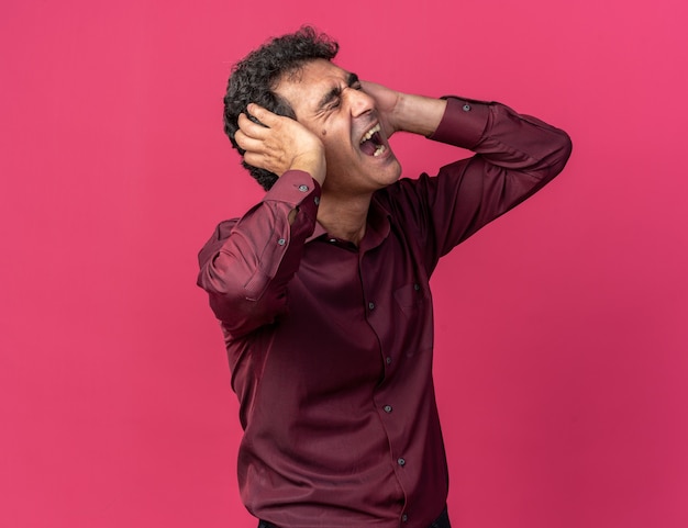 Senior man in purple shirt covering ears with hands shouting with annoyed expression 