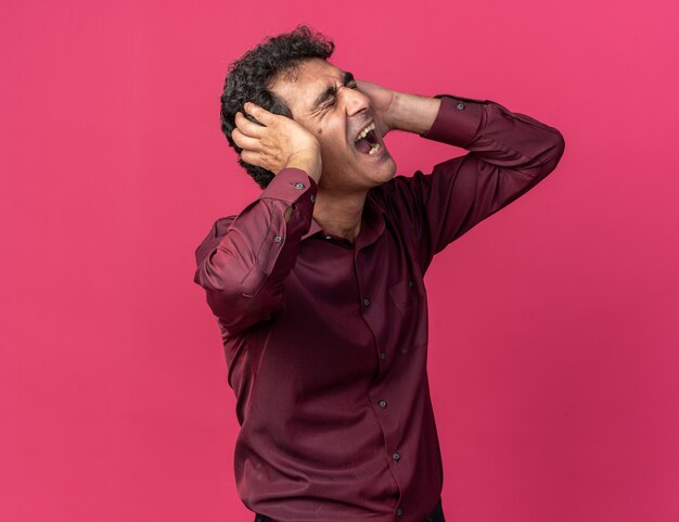 Senior man in purple shirt covering ears with hands shouting with annoyed expression 