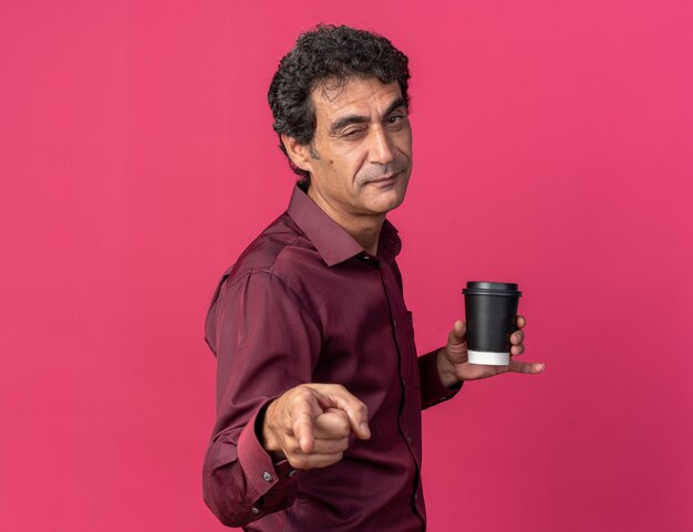 Senior man in purple shirt holding paper cup pointing with index finger at camera smiling and winking 