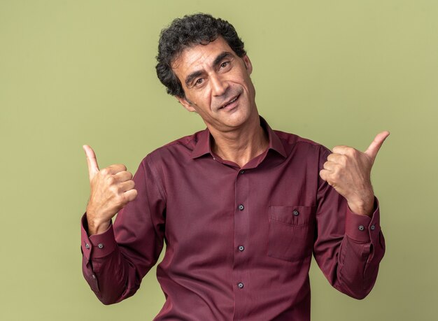 Senior man in purple shirt looking at camera happy and positive showing thumbs up standing over green