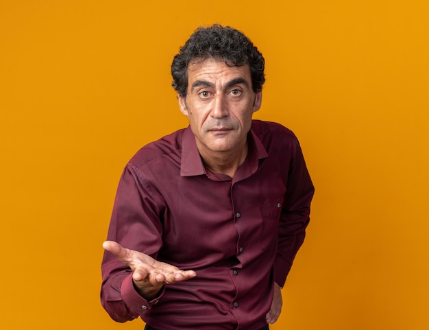 Free photo senior man in purple shirt looking at camera with arm out like asking a question standing over orange