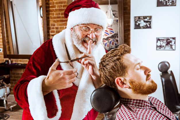 The senior man in Santa claus costume working as personal master with scissors at barber shop before Christmas