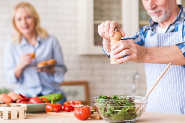 Senior man seasoning the green vegetables salad and her wife holding the muffins in hand at backdrop