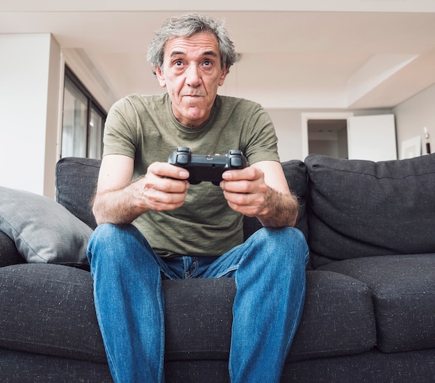 Senior man sitting on sofa playing video game with joystick