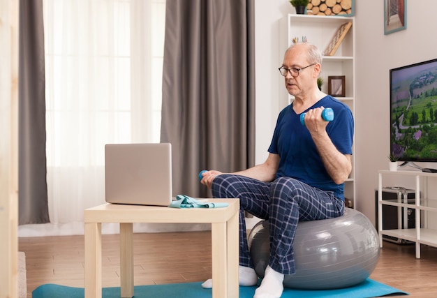 Senior man training with online coach sitting on swiss ball in the middle of the room