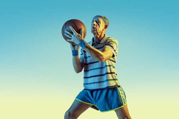 Senior man wearing sportwear playing basketball on gradient background, neon light. Caucasian male model in great shape stays active. Concept of sport, activity, movement, wellbeing, confidence.