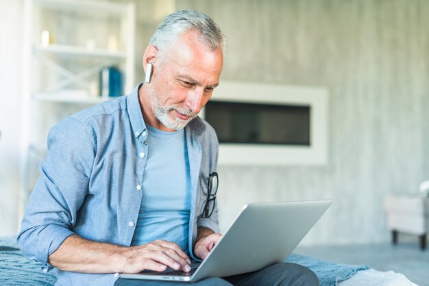 Senior man with wireless bluetooth sitting on bed using laptop