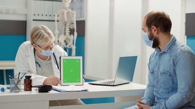 Free photo senior medic and patient using greenscreen on digital tablet during covid 19 pandemic. doctor showing blank mockup background with isolated copyspace chromakey on template to patient. tripod shot.