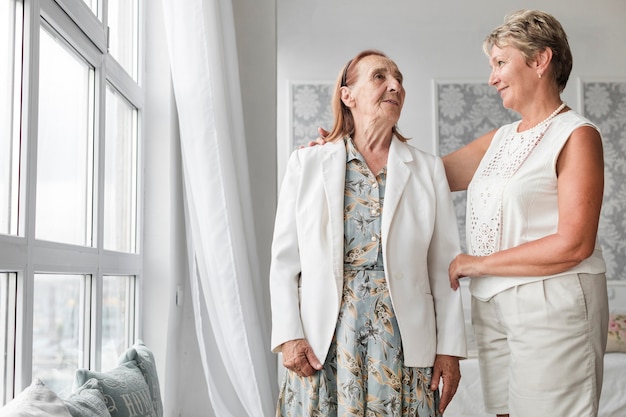 Senior mother and daughter looking at each other while standing near window