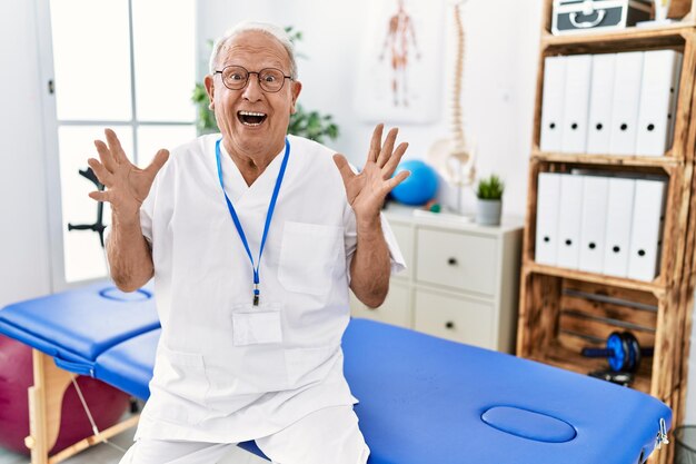 Senior physiotherapy man working at pain recovery clinic celebrating crazy and amazed for success with arms raised and open eyes screaming excited. winner concept