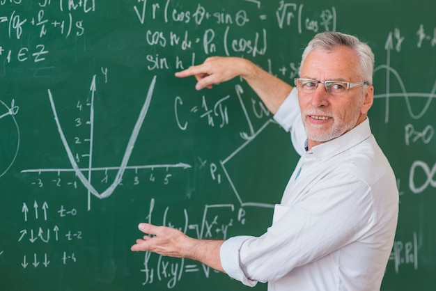 Senior teacher in glasses explaining math example on green chalkboard