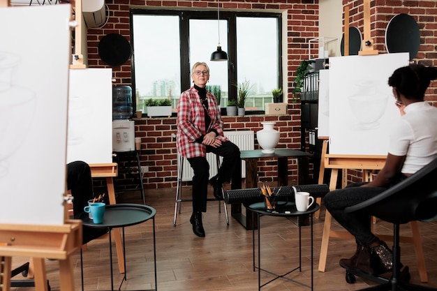 Free Photo senior woman artist teaching group of people how to draw, sitting on chair in classroom and smiling at camera, positive atmosphere at drawing workshop. art education and sketching classes for adults