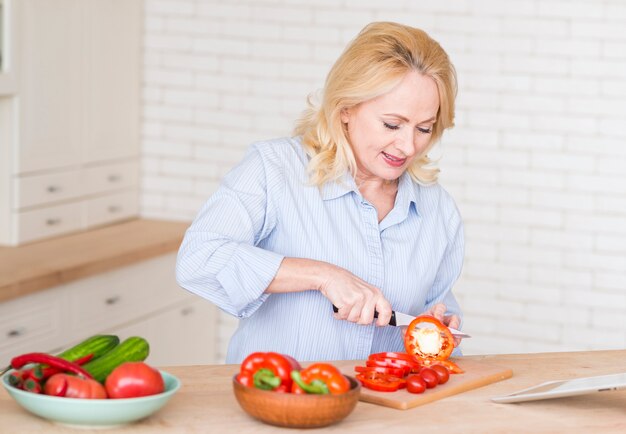 Free photo senior woman cutting the slices of red bell pepper with knife on chopping board in the kitchen