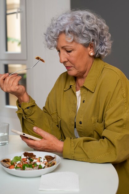 Senior woman eating figs dish at home and using smartphone