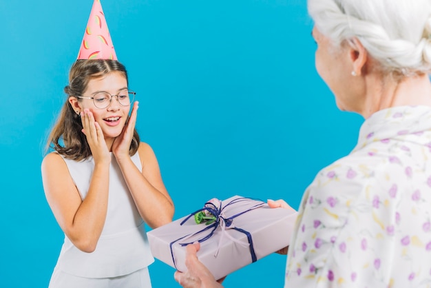 Senior woman giving birthday gift to her granddaughter on blue background