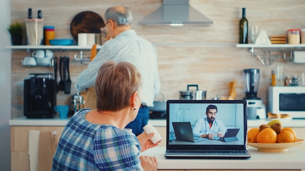 Free photo senior woman holding bottle of pills during video conference with doctor using laptop in kitchen. online health consultation for elderly people drugs ilness advice on symptoms, physician telemedicine