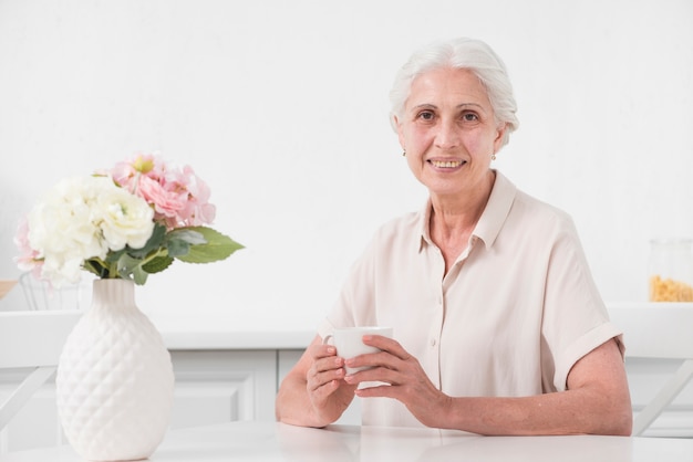 Senior woman holding cup of coffee with flower vase on white table