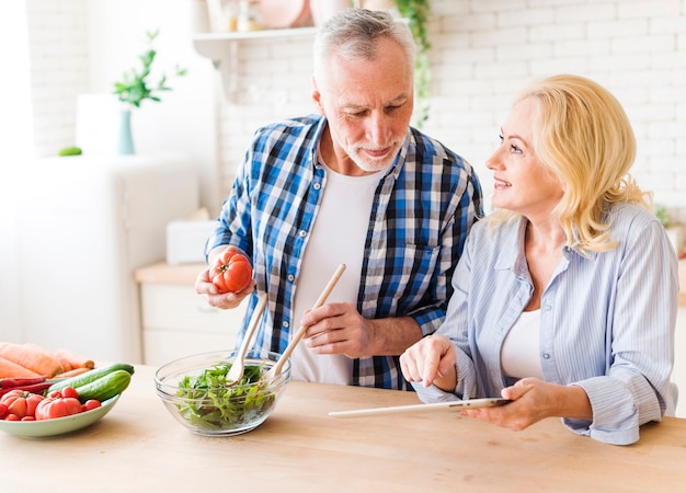 Senior woman holding digital tablet in hand showing recipe to her husband preparing the salad in the kitchen