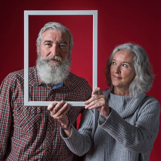 Senior woman holding white frame border in front of her husband's face against red backdrop