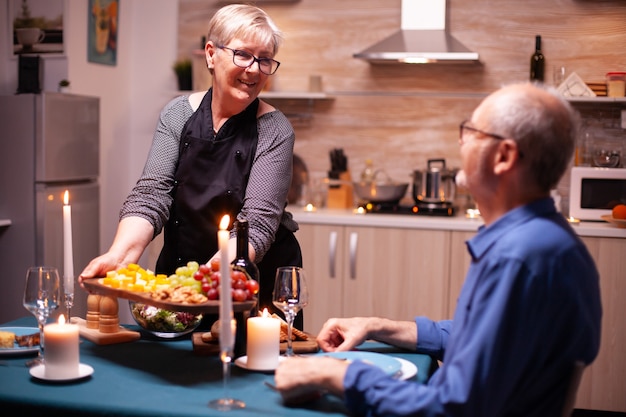 Senior woman holding wooden plate with and looking at husband during festive dinner. Elderly old couple talking, sitting at the table in kitchen, enjoying the meal, celebrating their anniversary .