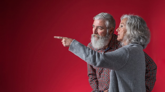 Senior woman showing something to her husband by pointing finger against red background