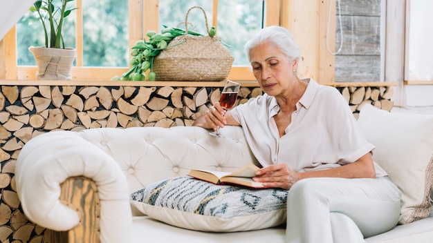 Senior woman sitting on sofa holding wine glass reading book