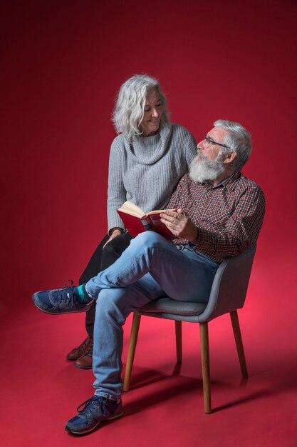 Senior woman sitting with her husband sitting on chair holding the book in hand against red background