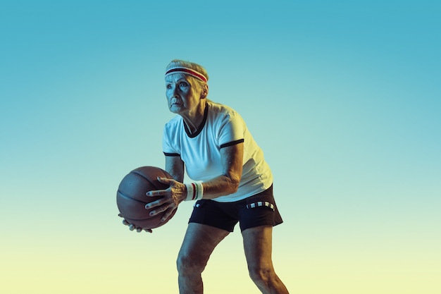 Senior woman in sportwear playing basketball on gradient background, neon light.