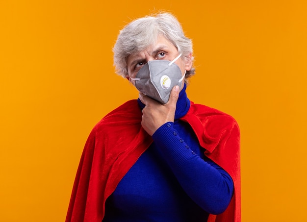 Free photo senior woman superhero wearing red cape and facial protective mask looking up puzzled standing over orange