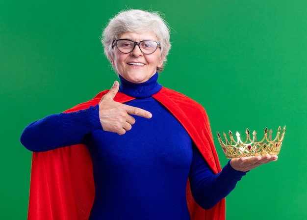 Senior woman superhero wearing red cape and glasses holding crown pointing with index finger at it smiling confident standing over green background