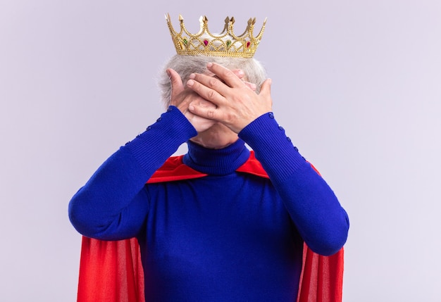 Free photo senior woman superhero wearing red cape and glasses with crown on head covering eyes with hands standing over white background