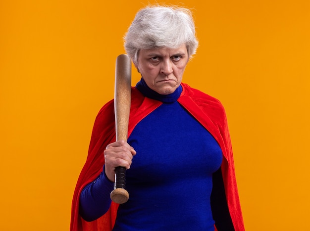 Free Photo senior woman superhero wearing red cape holding baseball bat looking at camera with serious face standing over orange background