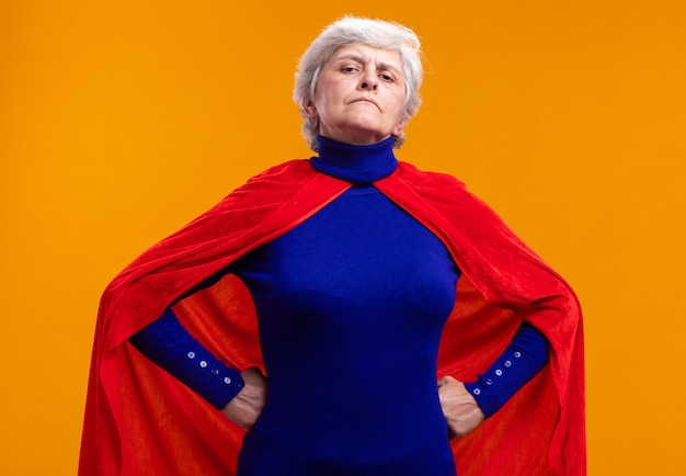 Free photo senior woman superhero wearing red cape looking at camera with confident expression with arms at hip standing over orange