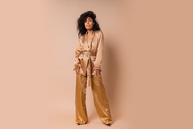 Sensual black woman with beautiful wavy hairs in elegant  golden satin suit posing over beige wall. Spring fashion look. Full lenght.