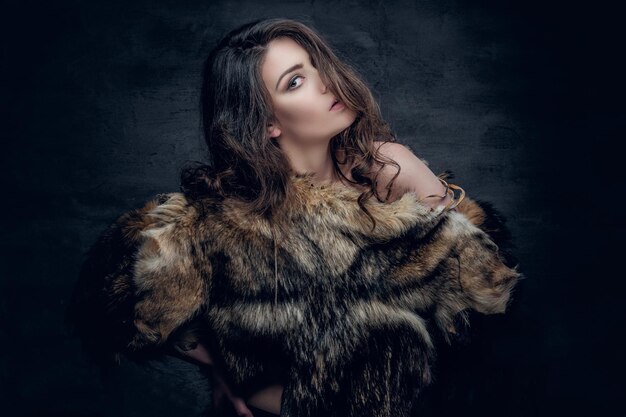 Sensual brunette female with curly hair dressed in a fur coat posing in deep shadow on grey background in a studio.