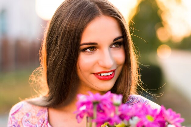 Sensual brunette woman posing with flower bouquet after romantic date, sunset colors, elegant dress and make up.