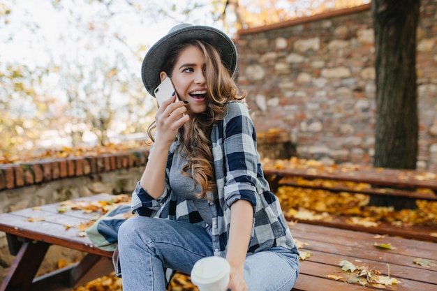 Sensual curly woman in hat expressing funny emotions during photoshoot in autumn yard