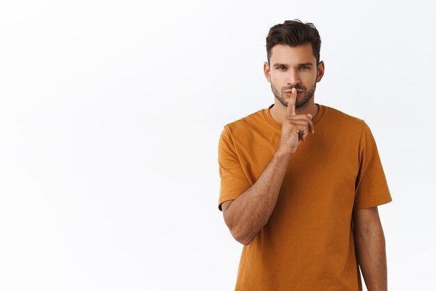 Sensual and dreamy handsome bearded man in brown tshirt shushing and squinting passionatly keep secret asking be quiet hushing romantically and mysteriously standing white background