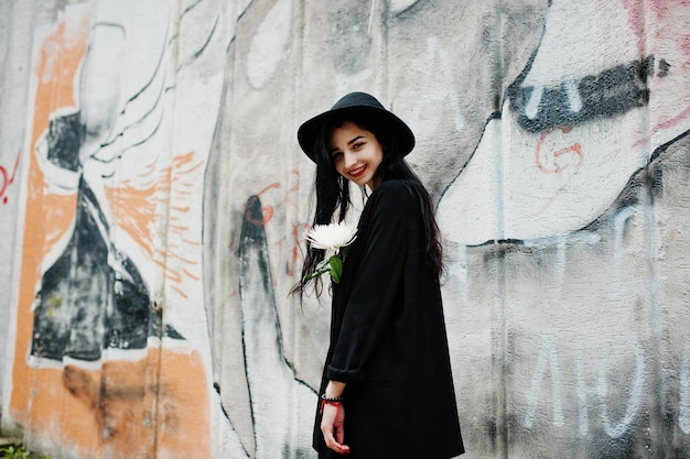 Free photo sensual girl all in black red lips and hat goth dramatic woman hold white chrysanthemum flower against graffiti wall