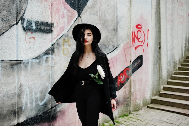 Free photo sensual girl all in black red lips and hat goth dramatic woman hold white chrysanthemum flower against graffiti wall