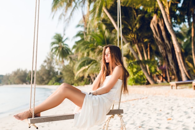 Free photo sensual tender girl sitting on a swing wearing white dress. girl has her eyes closed. she has long dark hair. she has bracelets on her arm and leg. the swing is on the beach with green palms