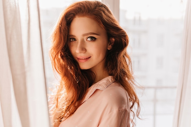 Sensual young woman with ginger hair standing near window. Indoor portrait of appealing white girl.