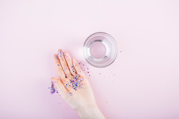 Sequins and glitter capsule on human hand near the glass of water against pink background