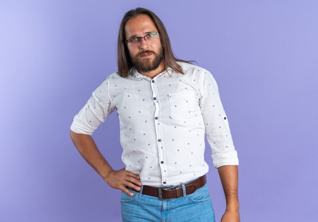 Free Photo serious adult handsome man wearing glasses keeping hand on waist looking at camera isolated on purple wall