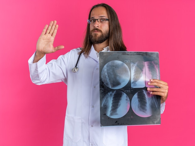 Free photo serious adult male doctor wearing medical robe and stethoscope with glasses showing x-ray shot looking at camera doing stop gesture isolated on pink wall