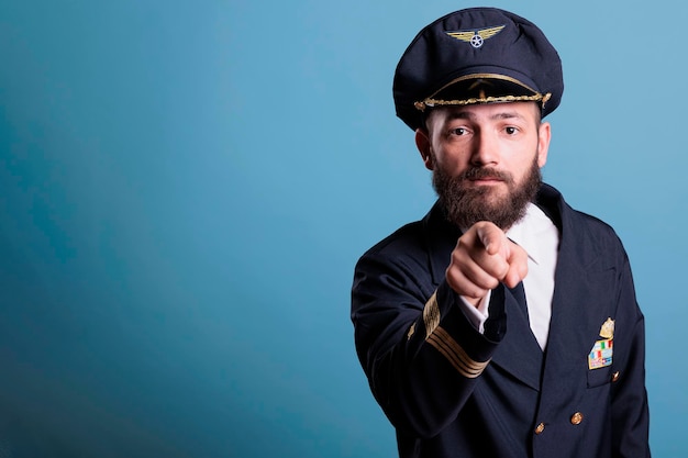 Free photo serious airplane pilot pointing at camera, aviation academy recruitment, plane captain wearing uniform and hat front view portrait. middle age aviator with airline wings badge on jacket