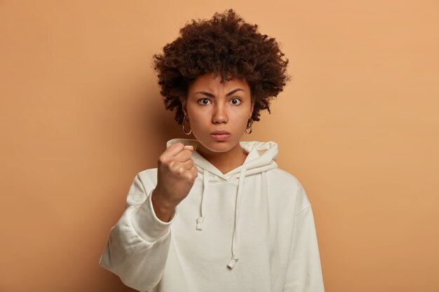 Serious angry ethnic woman threatens you with clenched fist, says I will show you how to disobey, wears casual white hoodie, promises to punish