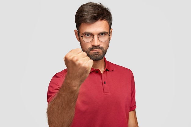 Serious angry male shows fist, ready for fight or challenge, has stern expression, wears casual red t-shirt, poses against white wall. Aggressive young man gestures indoor. Body language concept