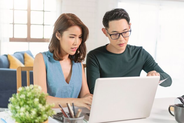 Serious Asian husband checking analyzing statement utilities bills sitting together at home.